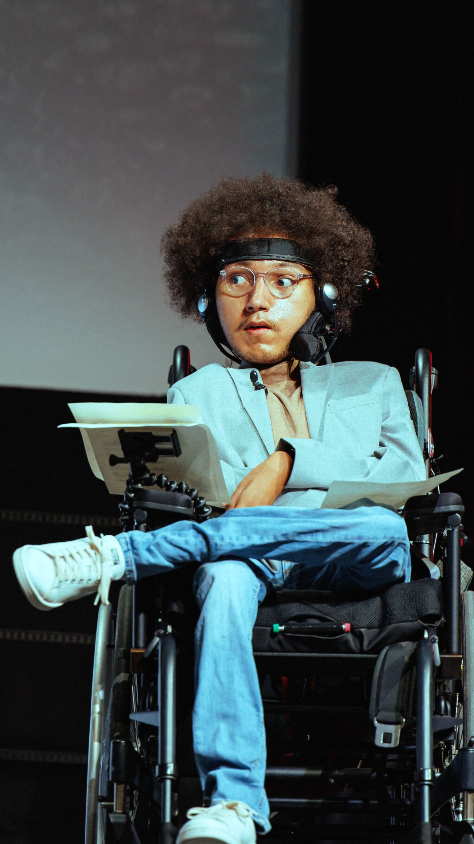 L'image montre Malick Reinhard assis dans un fauteuil roulant manuel. Il porte des vêtements décontractés chics, composés d'une veste de costume grise claire, un col roulé beige et un jean bleu. Il est chaussé de baskets blanches. Ses jambes son croisées. Le jeune homme a les cheveux crépus, porte des lunettes à monture ronde transparente et un casque-micro. Il semble concentré sur quelque chose hors de la vue, tenant des papiers ou un livret. L'arrière-plan est flou avec une teinte de gris, suggérant un éclairage de scène ou une présentation.