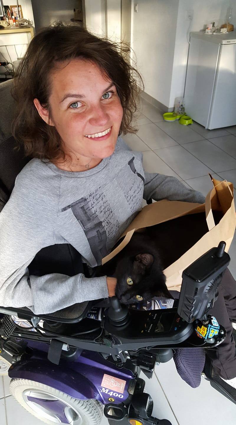 Une femme souriante en fauteuil roulant, Pamela Ruga, vêtue d'un pull gris, tient un sac en papier brun contenant un chat noir. La scène se déroule dans une cuisine, avec des bols pour animaux au sol en arrière-plan.