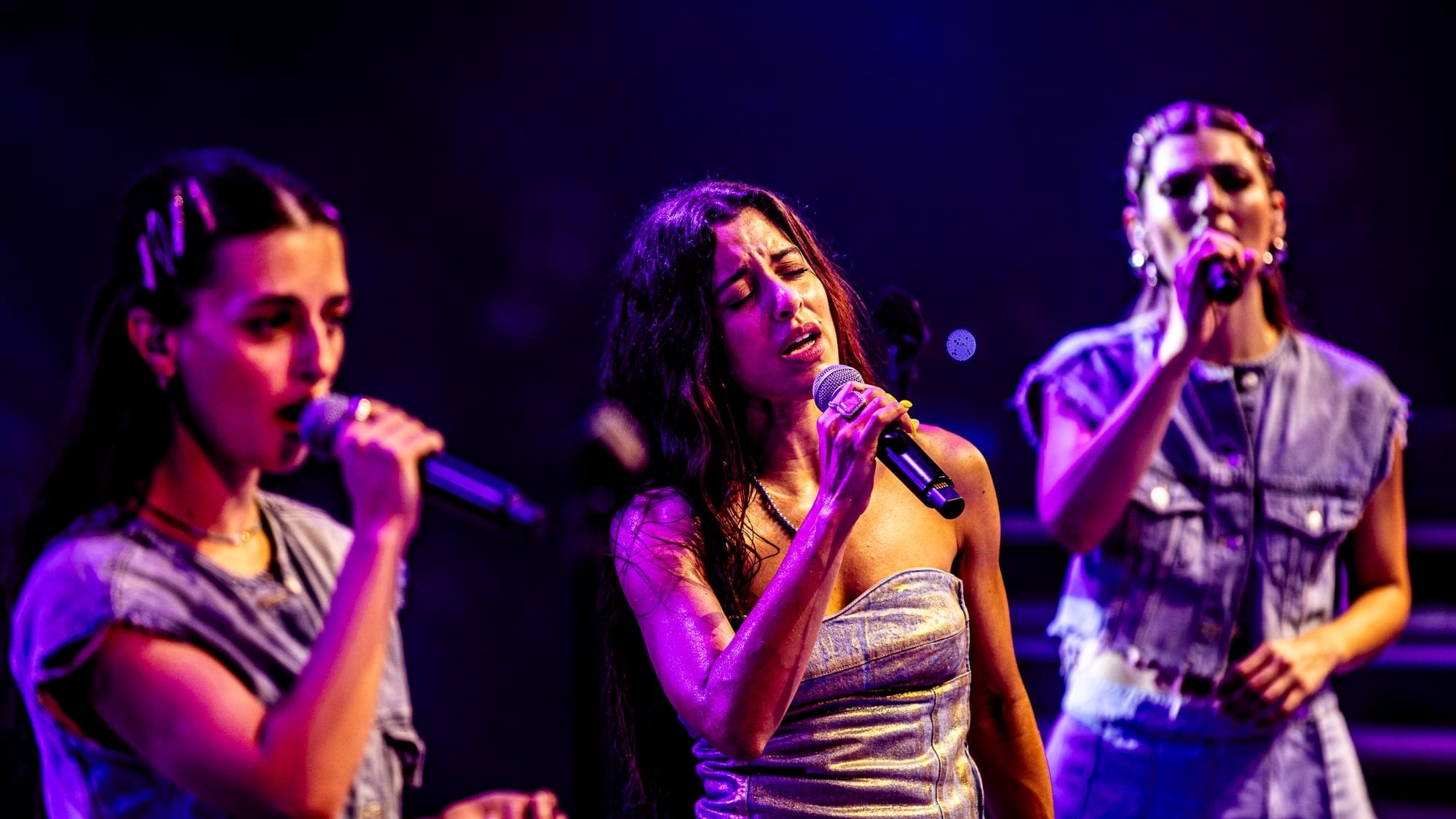 Marina Satti et ses choristes sur scène au Paléo Festival Nyon 2024. Les trois chanteuses interprètent avec passion, chacune tenant un micro. Marina Satti, au centre, porte une robe argentée et chante les yeux fermés, exprimant une intense émotion. Les choristes de chaque côté contribuent à l'harmonie vocale, créant une performance captivante sous des lumières violettes et bleues.