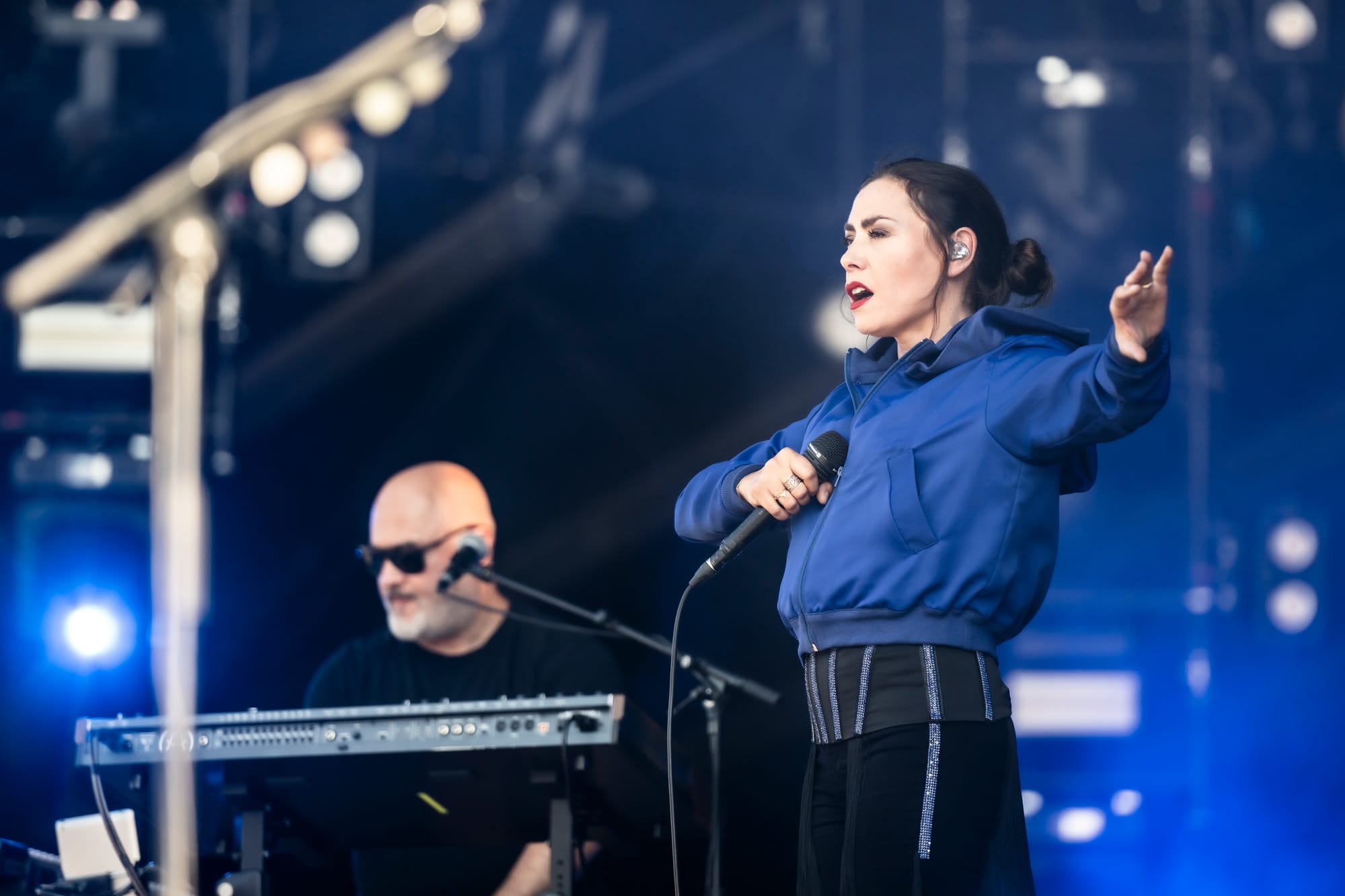 Olivia Ruiz, vêtue d'une veste bleue, chante avec passion sur scène au Paléo Festival 2024. Un musicien au clavier l'accompagne sous les projecteurs.