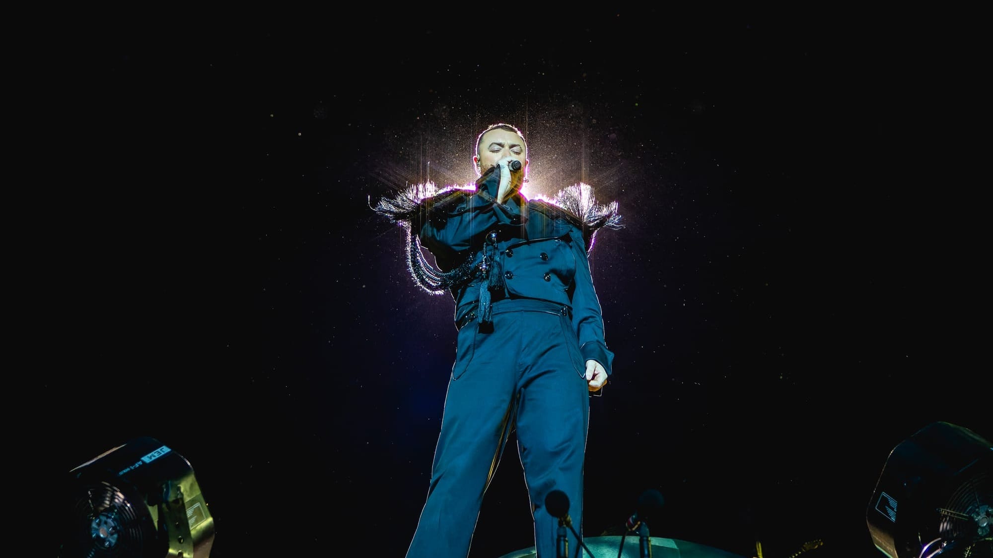 Sam Smith, vêtu d'un ensemble bleu électrique, chante passionnément sur scène, entouré d'effets lumineux créant une aura magique.