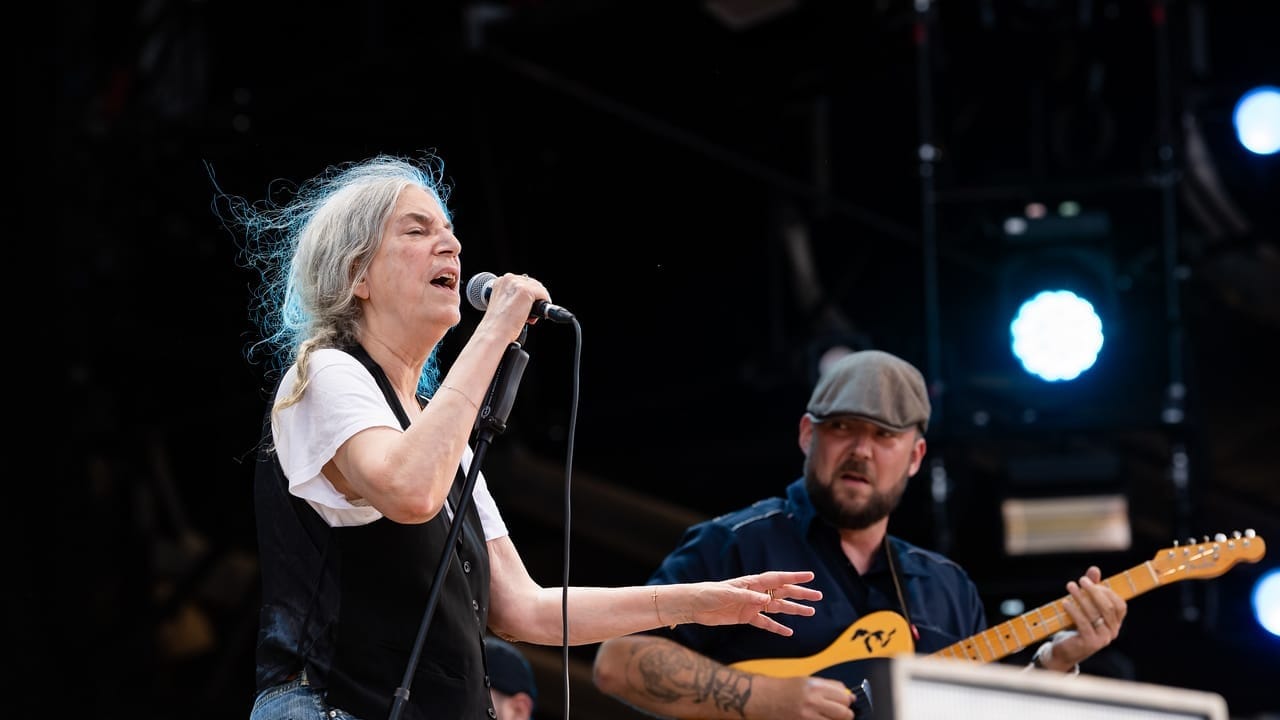 Patti Smith, vêtue d'un jean et d'un gilet noir, chante au micro sur scène lors du Paléo Festival de Nyon 2024, accompagnée d'un guitariste portant une casquette et jouant de la guitare électrique.