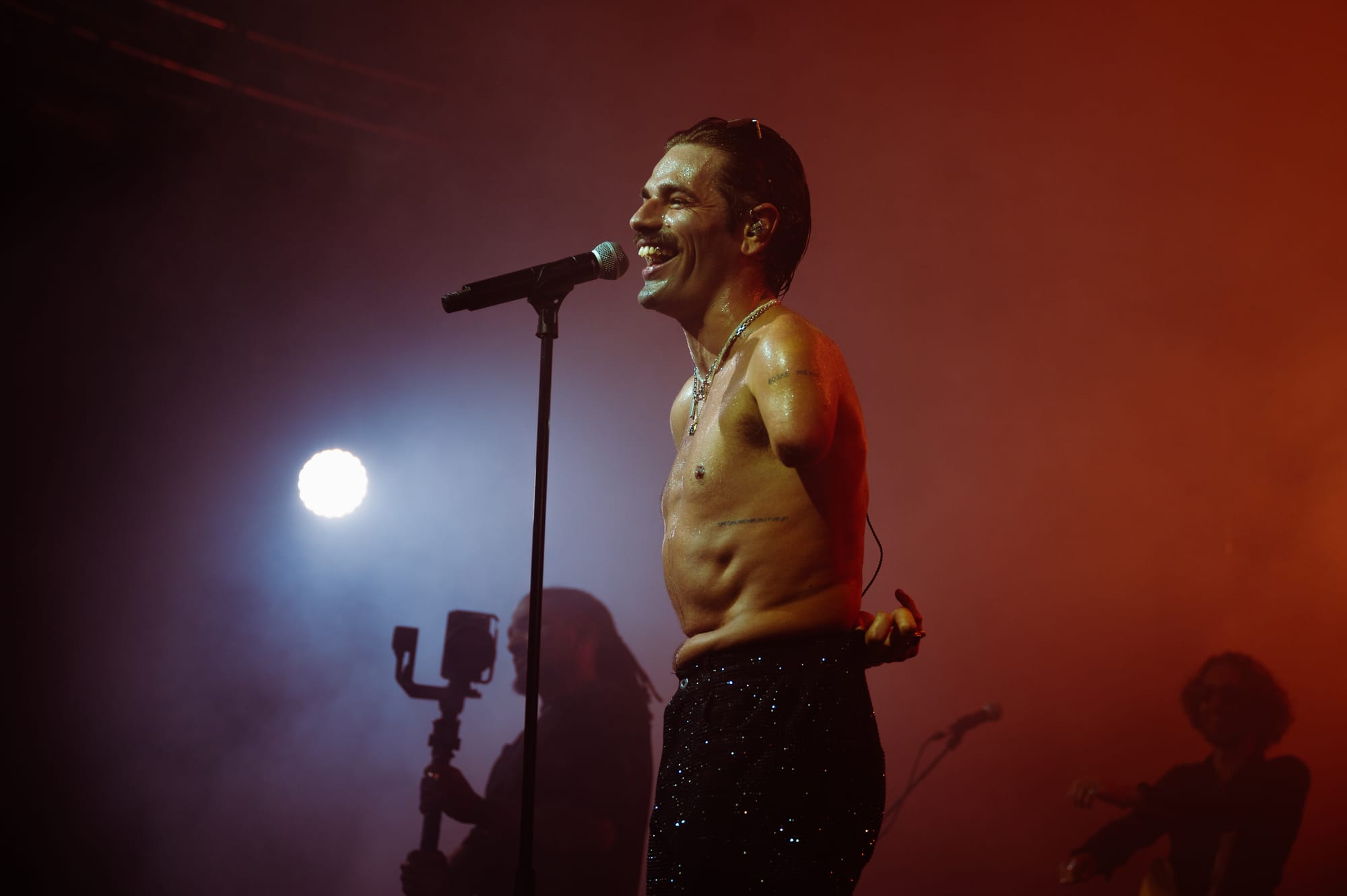 Lucky Love, souriant et torse nu, chante avec passion sur la scène du Paléo Festival 2024. Un éclairage rouge et bleu crée une ambiance électrisante. Il présente un bras gauche amputé.