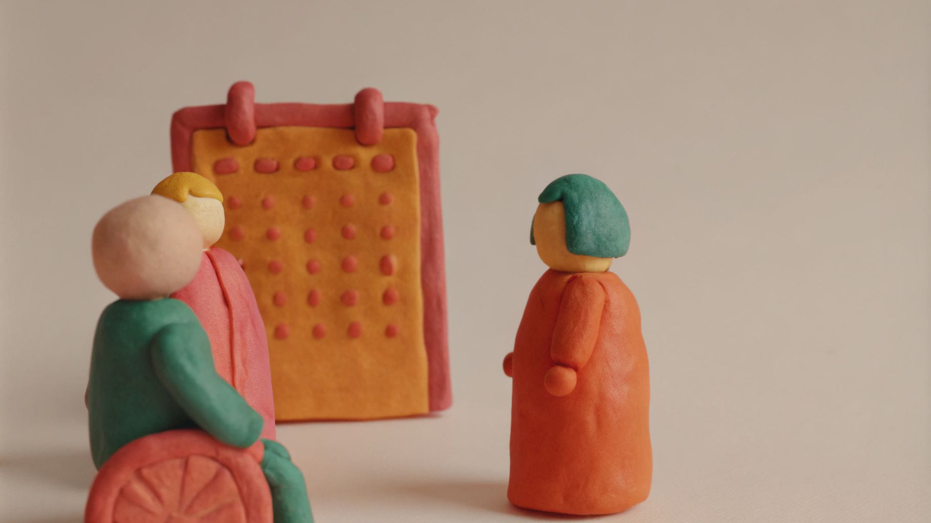 Scène en pâte à modeler représentant une personne en fauteuil roulant face à un calendrier mural orange avec des points rouges. La personne porte un vêtement vert et rose. En face d'elle se tient une autre figurine, debout, vêtue d'orange avec des cheveux verts.