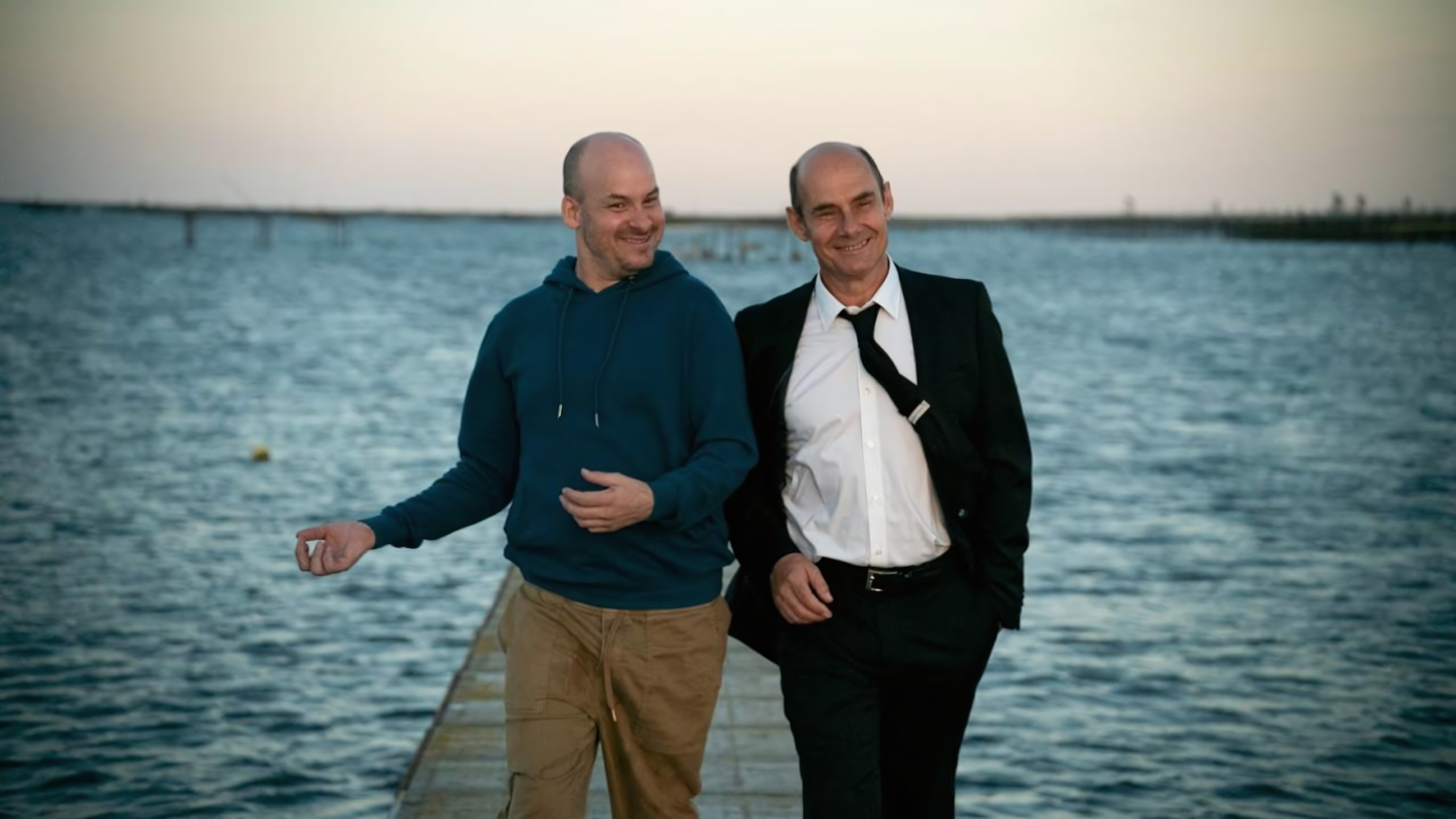 Sur un ponton au bord de l'eau au crépuscule, Alexandre Jollien (à gauche, en sweat à capuche bleu marine et pantalon beige) et Bernard Campan (en costume noir et chemise blanche, cravate légèrement défaite) se tiennent côte à côte, partageant un moment de complicité souriante. L'arrière-plan montre une étendue d'eau calme sous un ciel doux de fin de journée. 